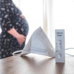 A mask and covid test in the foreground while a pregnant woman stands in the background
